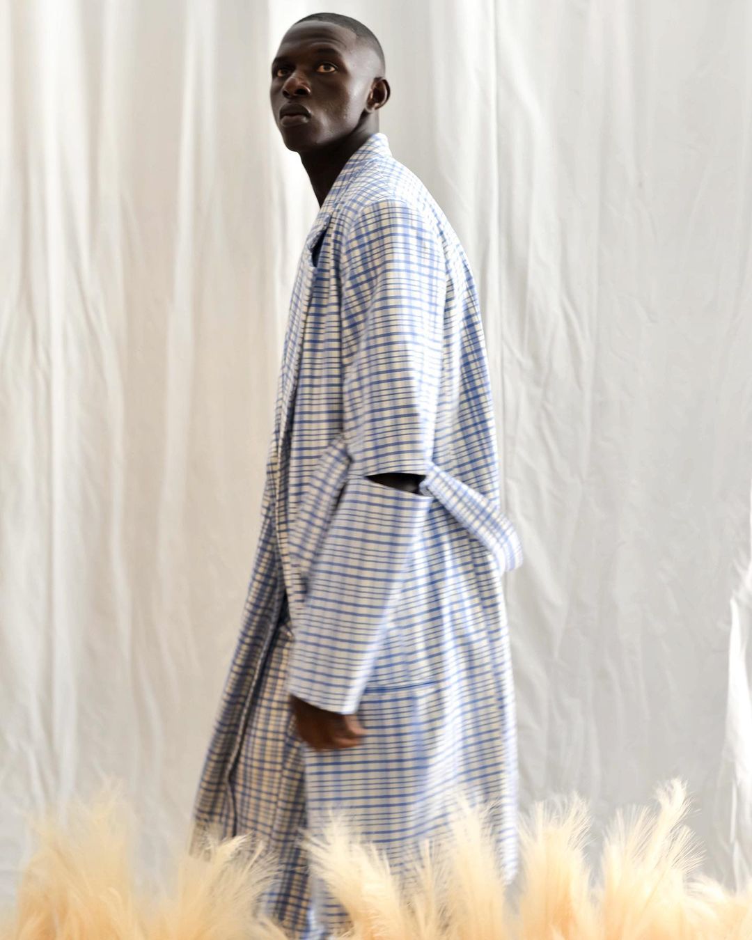 Fashionable African male model in a blue and white plaid robe poses in a minimalist setting with decorative pampas grass.