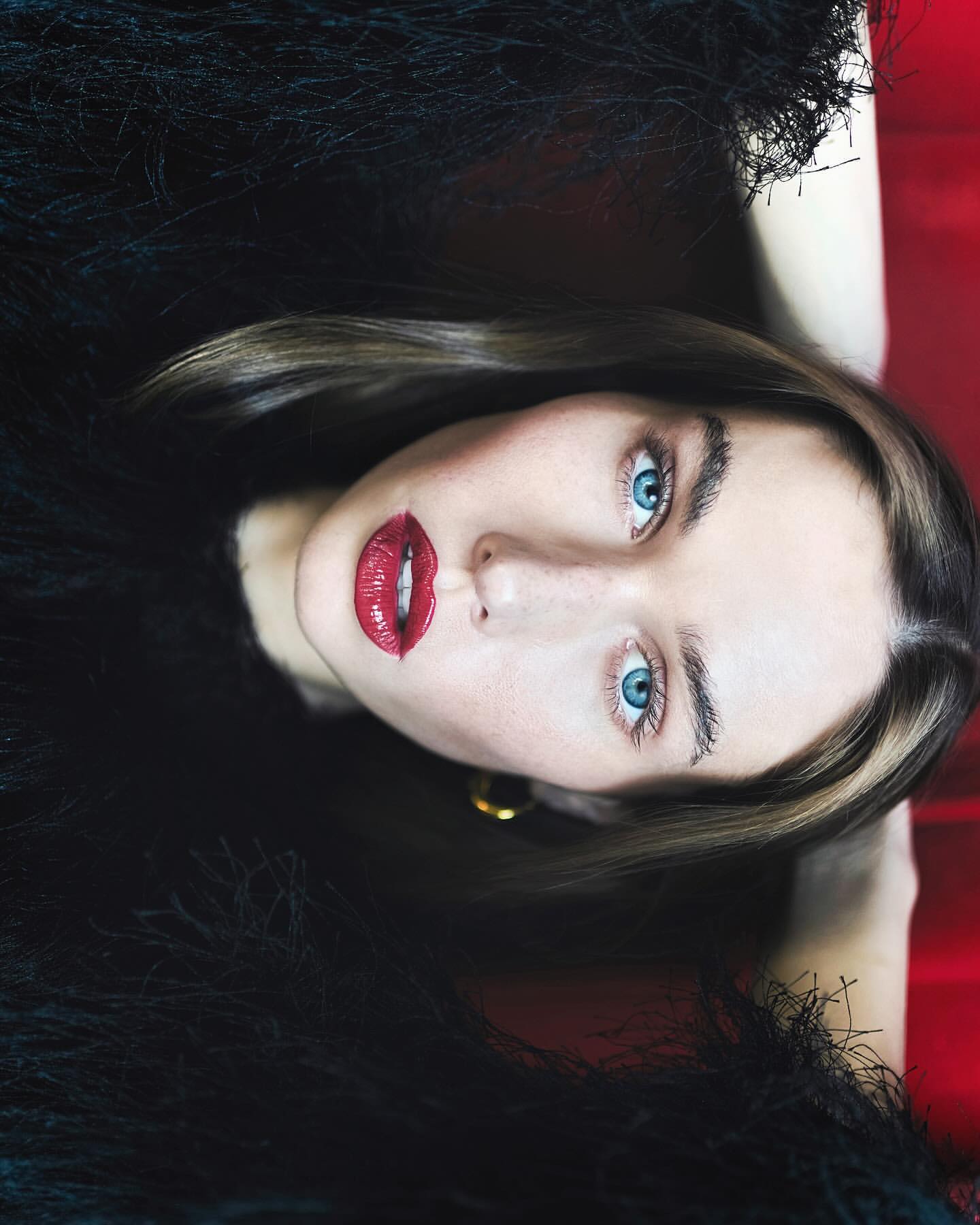 Close-up portrait of a woman with blue eyes and red lipstick wearing a feathered top, set against a red background.