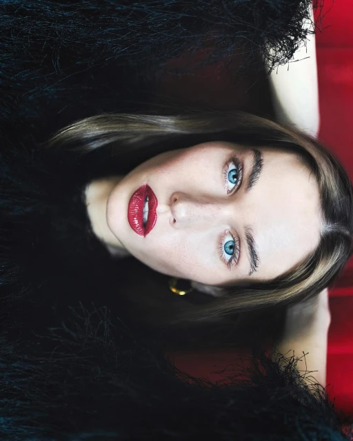 Close-up portrait of a woman with blue eyes and red lipstick wearing a feathered top, set against a red background.