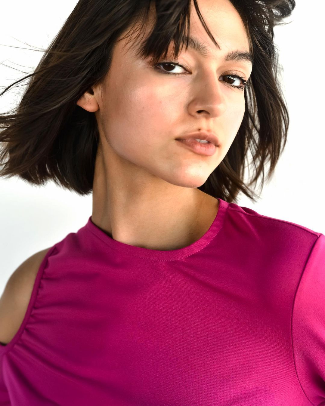 Portrait of a young woman with dark hair wearing a vibrant pink top against a white background, looking directly at the camera.