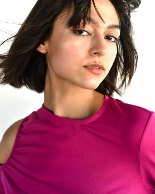 Portrait of a young woman with dark hair wearing a vibrant pink top against a white background, looking directly at the camera.