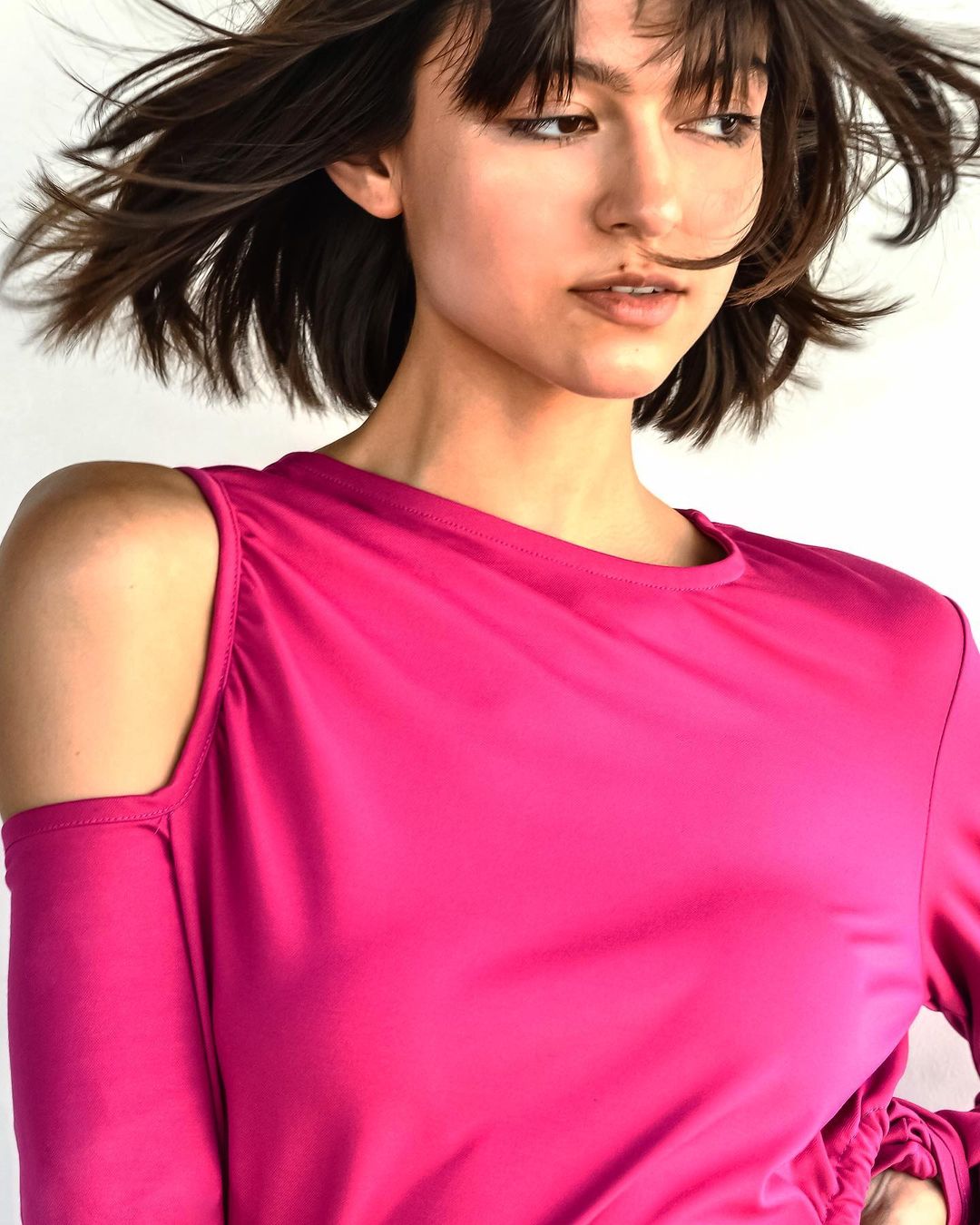 A woman with short brown hair poses in a pink long-sleeve, cold-shoulder top against a plain white background in a close-up shot.