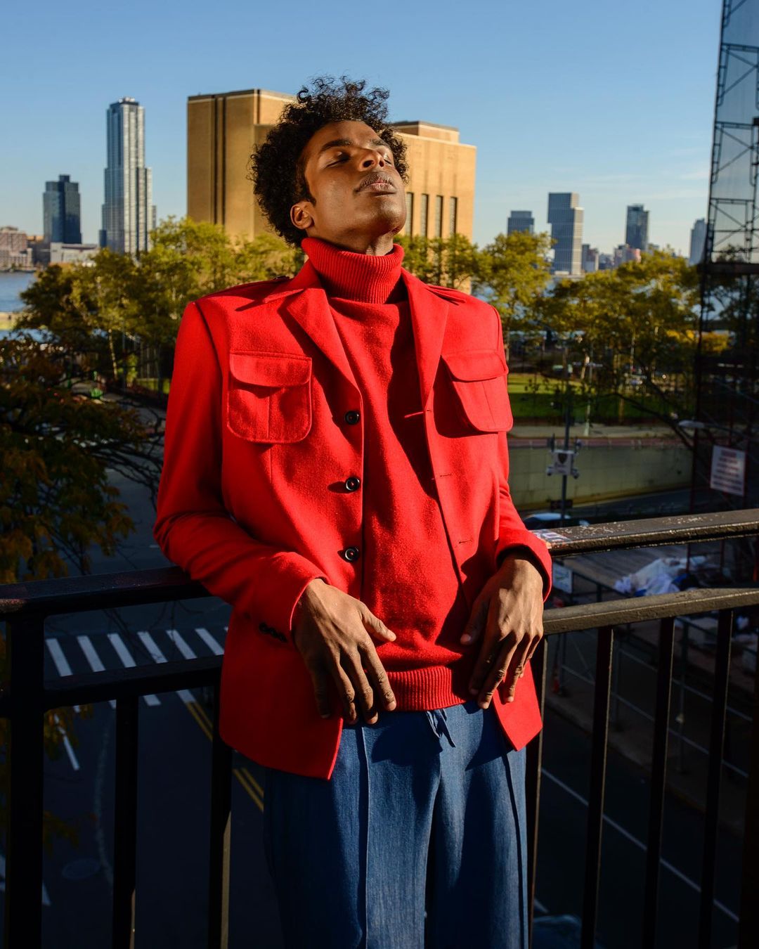 Adult male in a red jacket and blue pants stands on a balcony with his face to the sky, framed by a cityscape in the background.