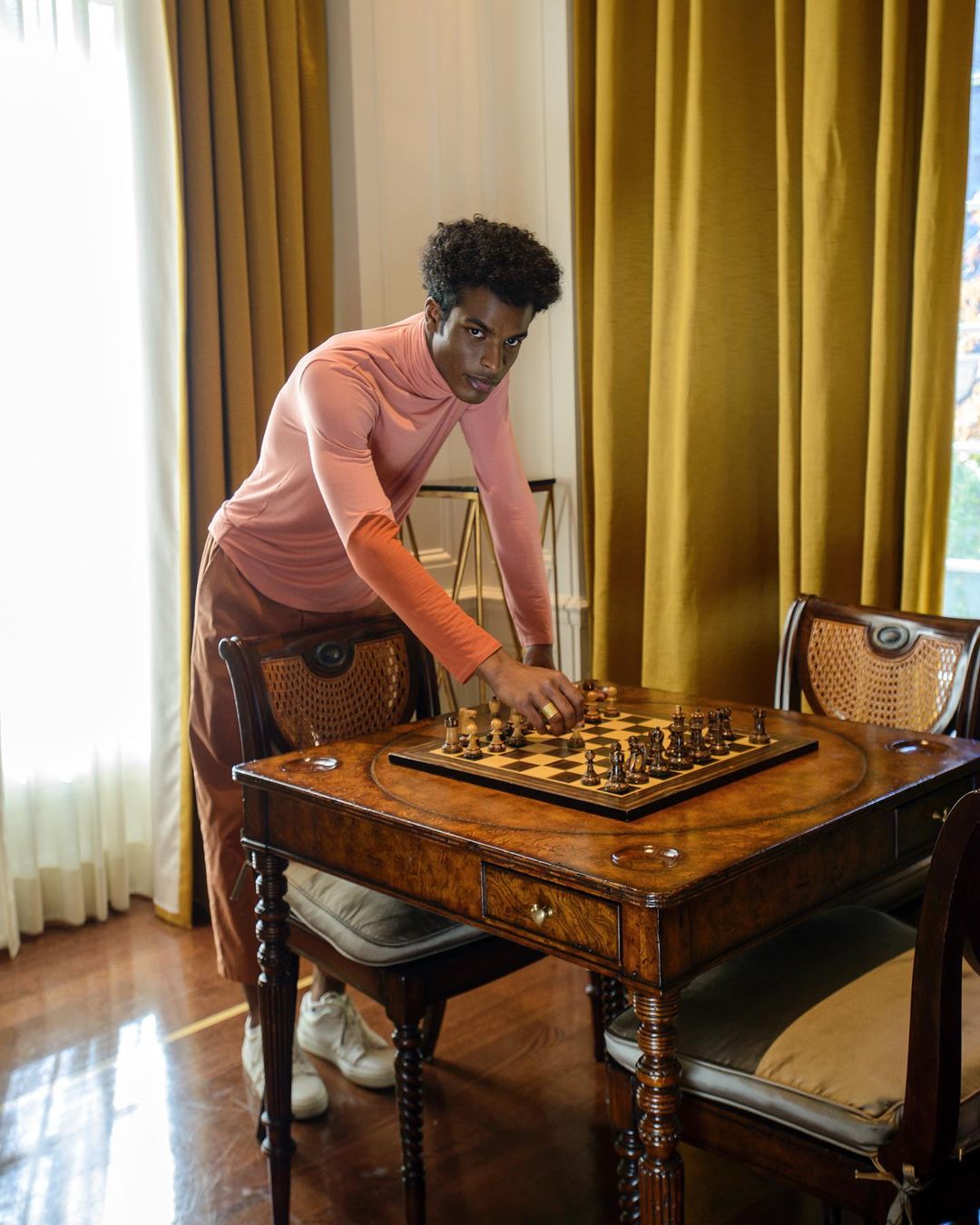 A young, adult man in an apricot turtleneck playing chess on a dark wood table in a naturally lit room with mustard-yellow curtains.
