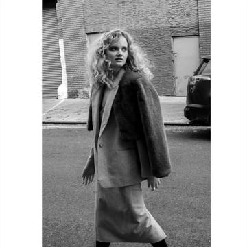 Black and white outdoor shot of a woman with curly hair wearing a jacket and skirt on a street with brick buildings.