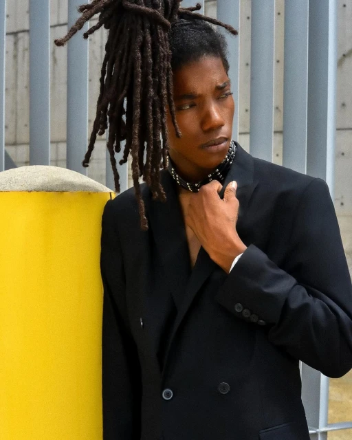 Portrait of an adult with dreadlocks wearing a black suit and studded choker, posing in front of a gray metal wall with a yellow column.