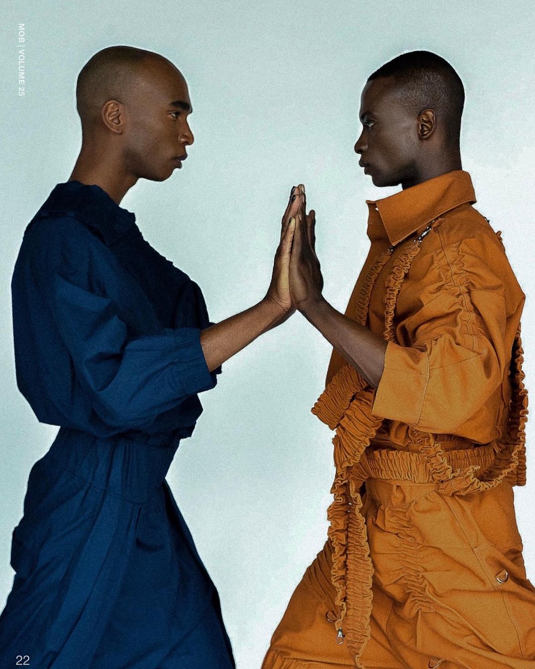 Two Black male fashion models in blue and orange outfits face each other with hands touching in a studio setting.