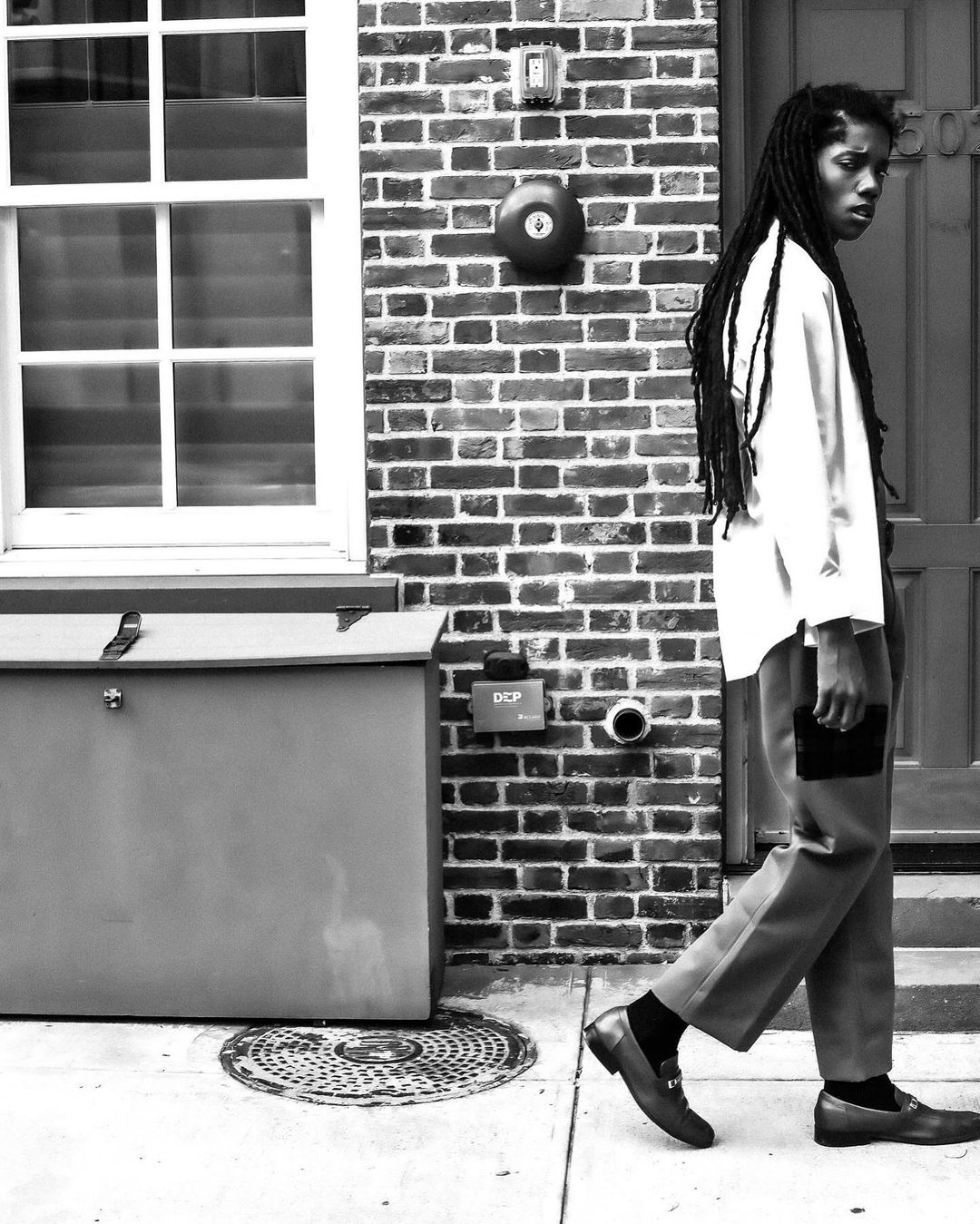 Monochrome outdoor shot of a person with dreadlocks in stylish trousers and shirt, walking by a brick wall and metal utility box.