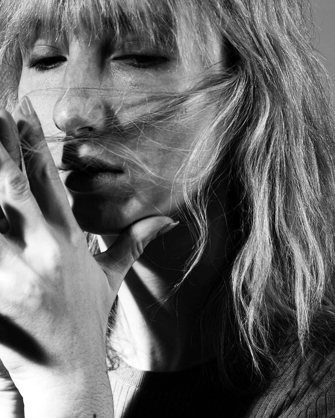 Monochrome portrait of a woman with blonde wavy hair, partially covering her face, her hand raised near her face in soft light.