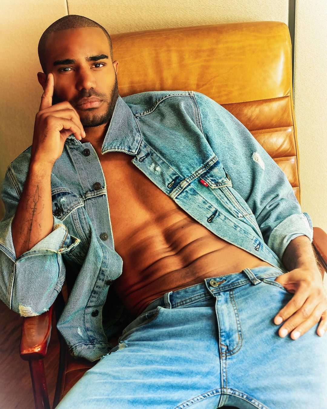 Shirtless black man wearing denim jacket and jeans seated in a brown leather chair, photographed in Milan.