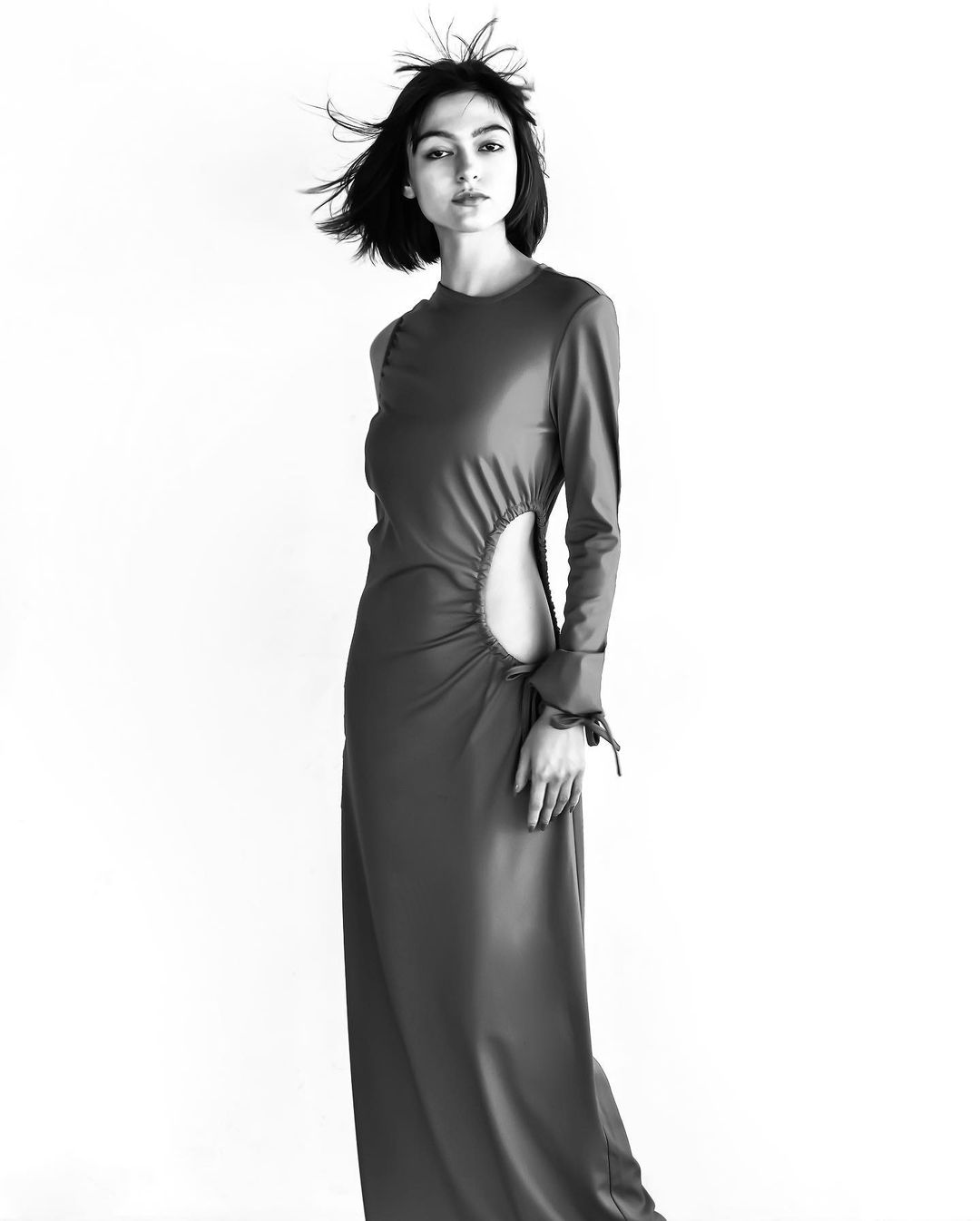 Black and white shot of a woman in a long dress with a cutout, posing in a studio with her hair blowing for a fashion editorial.