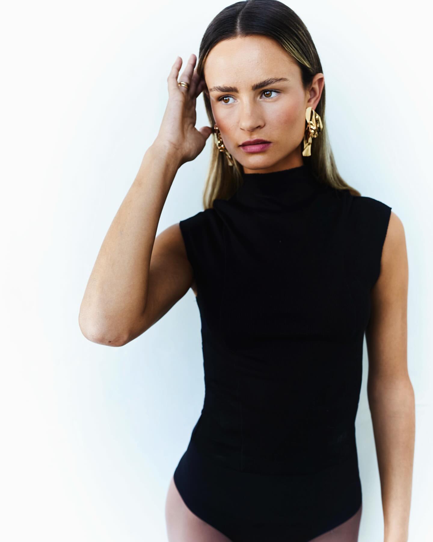 A woman with long blonde hair is posing in a black bodysuit and gold earrings against a light neutral background in a medium shot.