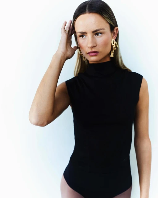 A woman with long blonde hair is posing in a black bodysuit and gold earrings against a light neutral background in a medium shot.
