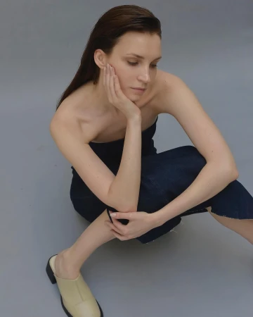 Model with dark wet-look hair sitting on a grey floor wearing a strapless dark denim dress and beige mules
