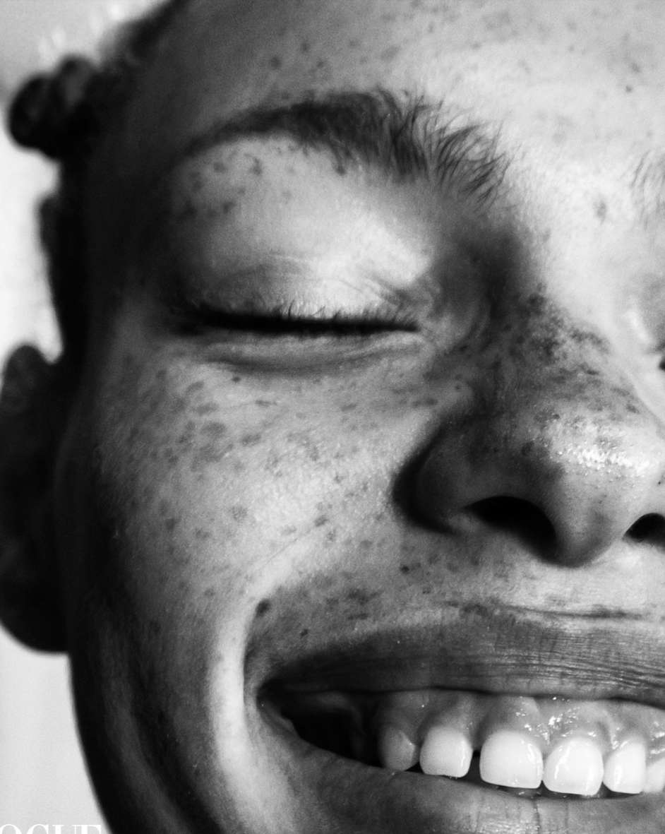 Black and white close-up of a smiling Black woman's face with freckles, eyes closed, and visible teeth, cropped at 'VOGUE' magazine.