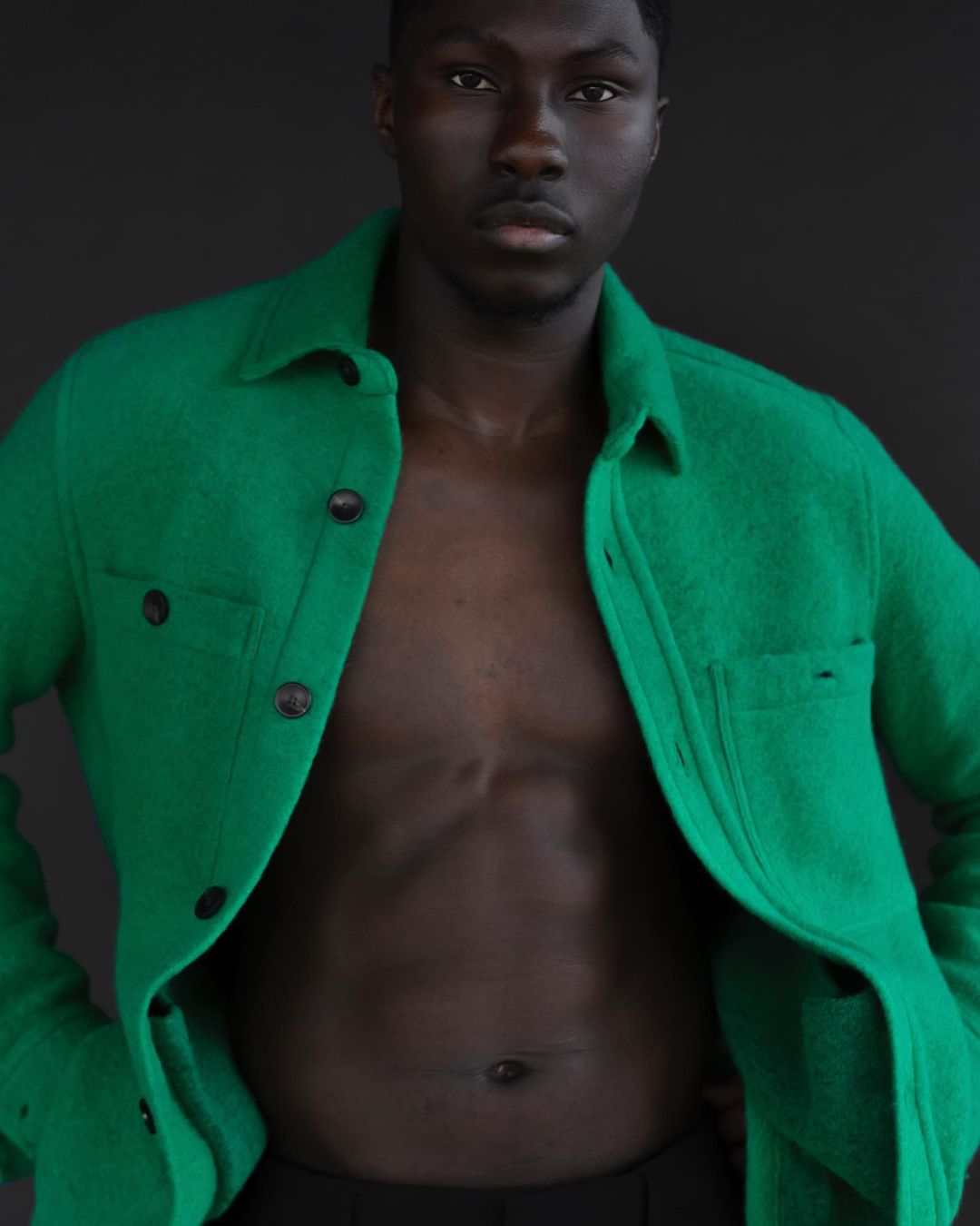 Dark-skinned male poses indoors wearing a green button-down shirt; portrait shot against a dark background. Fashion advertisement.