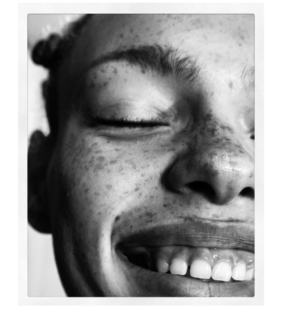Close-up black and white portrait of a smiling adult woman with freckles and eyes closed in a square white frame.
