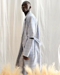 Black man wearing a blue and white plaid coat standing in front of a draped white textile background.