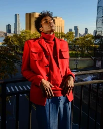 A young Black man wearing a vibrant red jacket and turtleneck leaning against a railing with city skyscrapers in the background.