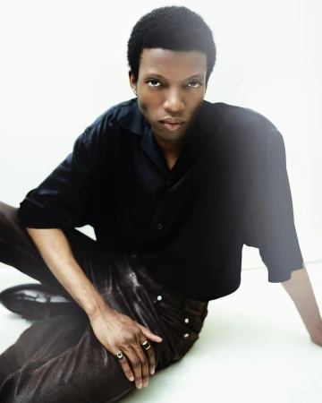 A Black man wearing a black shirt and dark trousers seated against a bright white background, photographed by Celestia Seyvant.