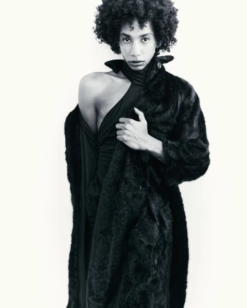 Black-and-white portrait of a model with voluminous curly hair wearing a textured dark coat draped over one shoulder against a white wall.