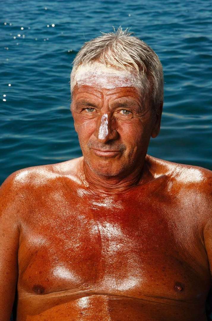 Older man with tan skin and sunscreen on his forehead and nose is sitting in a body of water, portrait orientation.