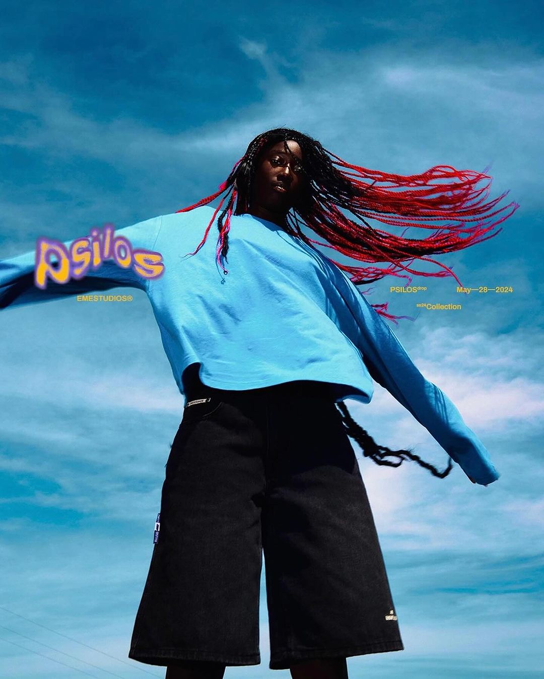 Woman in an oversized blue sweatshirt and black denim shorts against a bright blue sky with light clouds, fashion editorial.