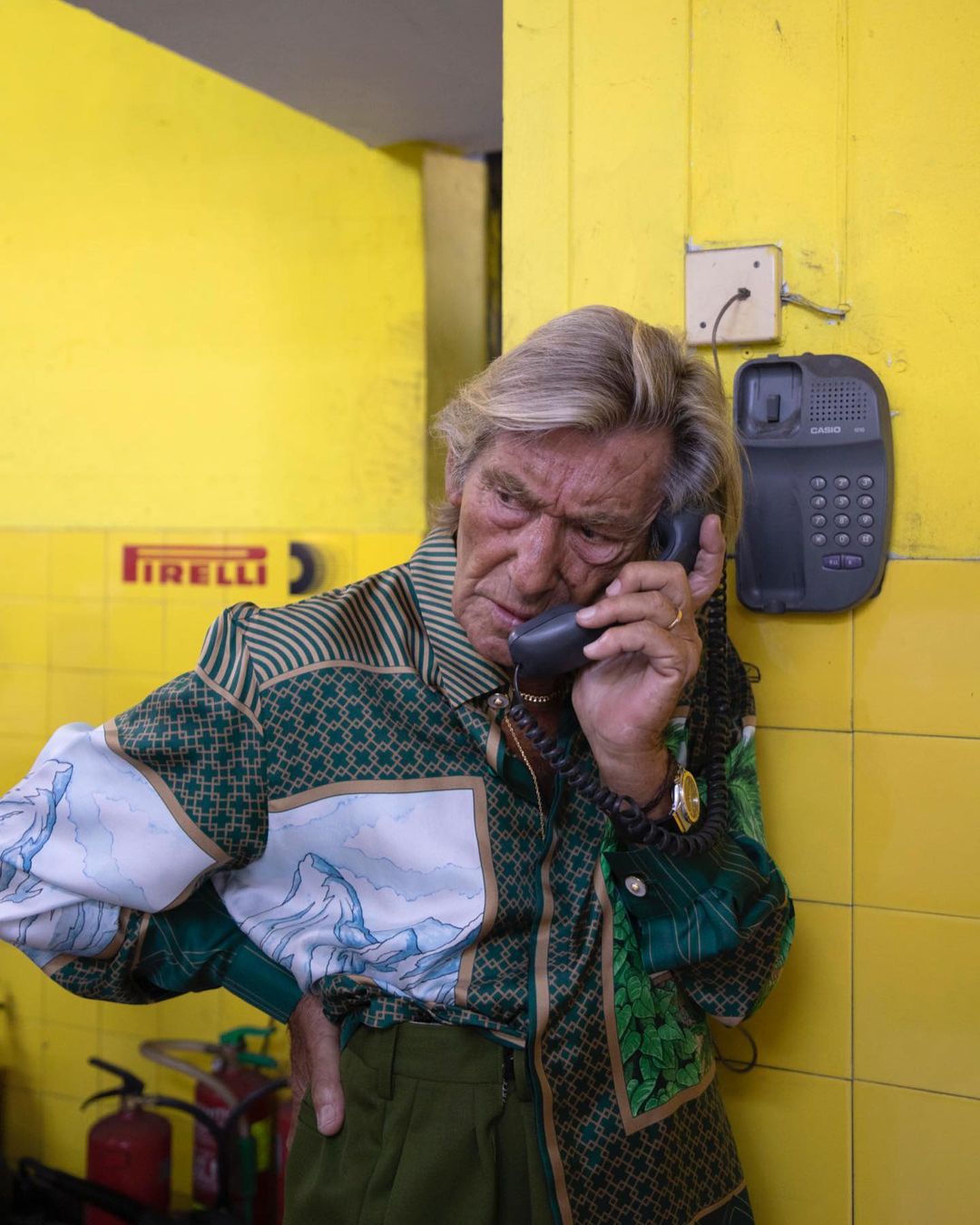 Mature adult on landline phone against yellow wall; wearing patterned collared shirt. Indoor shot with visible landline phone.