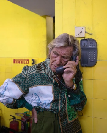 Mature adult on landline phone against yellow wall; wearing patterned collared shirt. Indoor shot with visible landline phone.