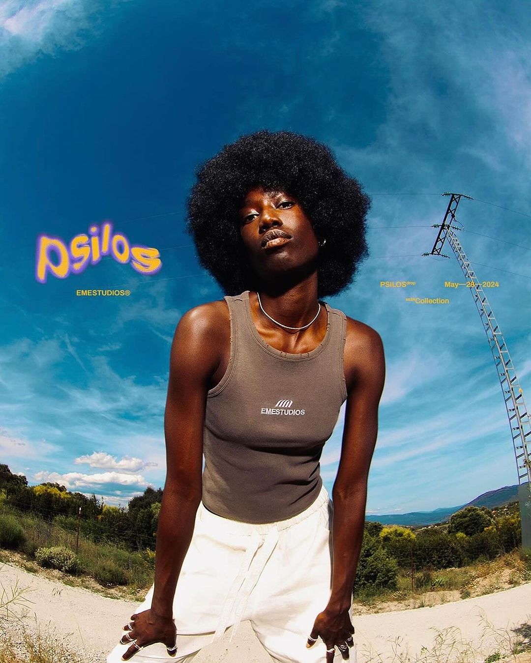 Outdoor fashion portrait of a Black woman with an afro wearing a tank top and white pants, with a rural landscape and blue sky background.