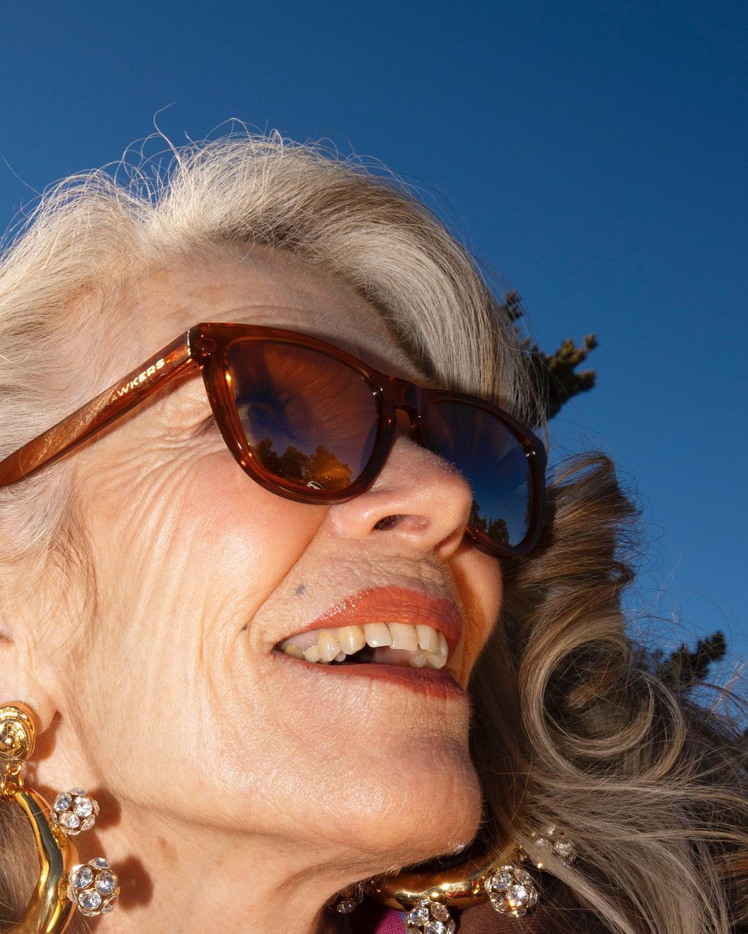 Close-up of a smiling mature woman with sunglasses and jewelry against a clear blue sky, capturing a sunny and fashionable portrait.