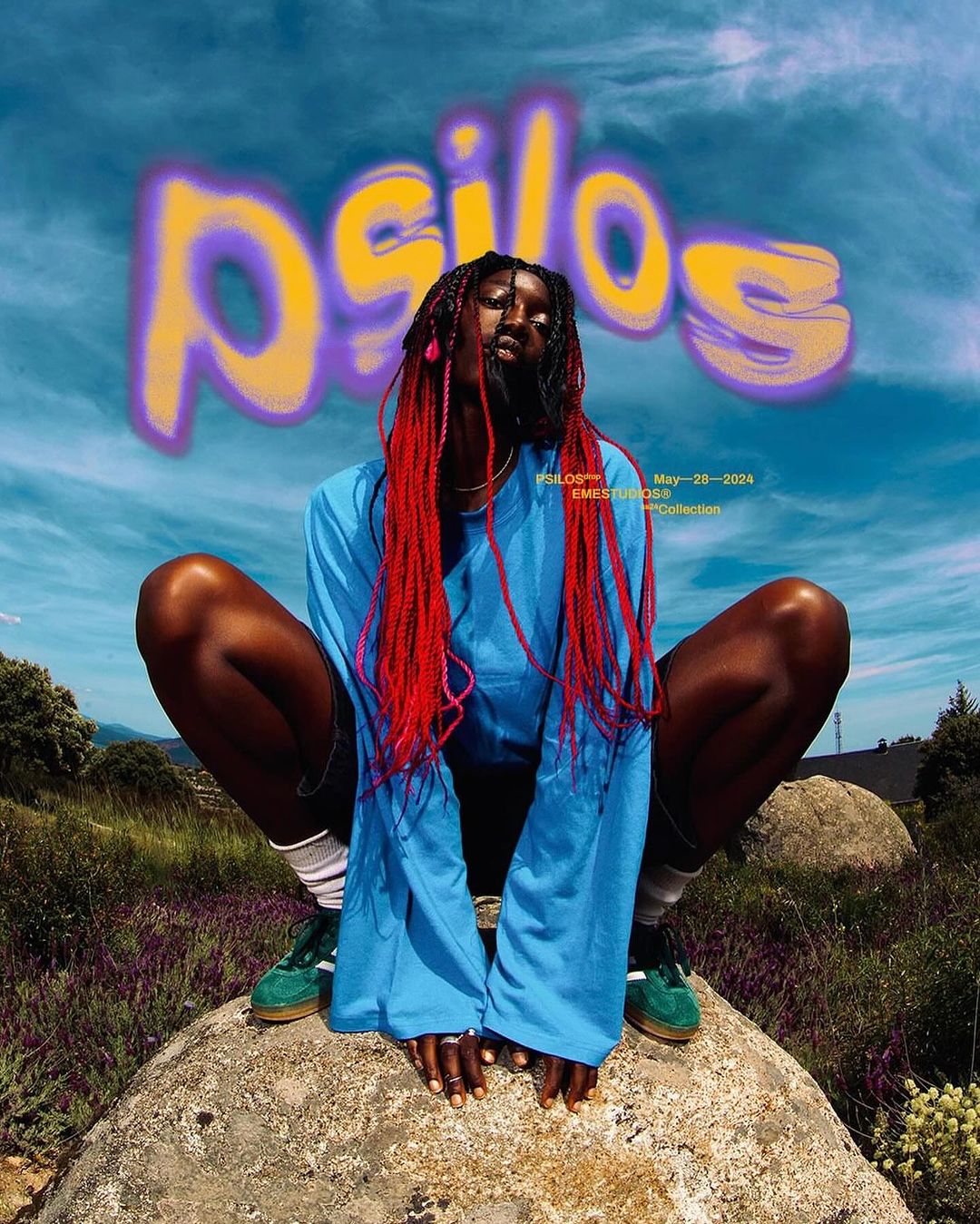 Portrait of a Black woman with red braids posing on a rock outdoors, wearing a long-sleeved blue shirt and green shoes against a cloudy sky.