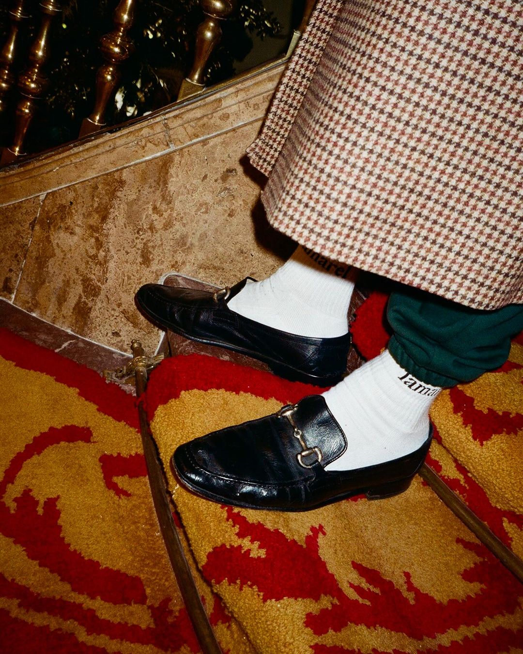 Close-up on black leather loafers worn with white socks on patterned carpet and marble stairs, featuring brown houndstooth skirt.