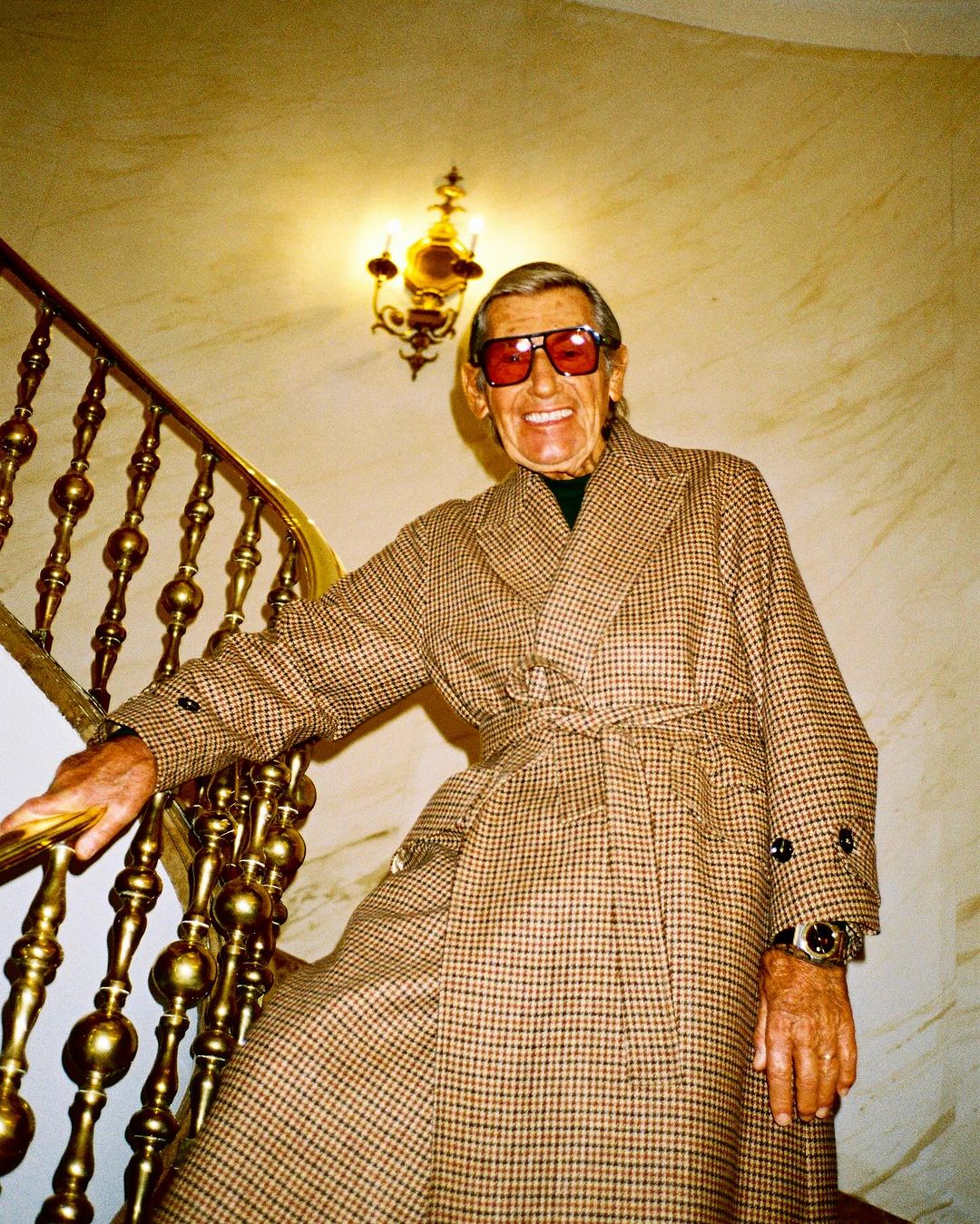 Elderly man in a checkered overcoat poses on a staircase with gold railings, lit by a chandelier, wearing sunglasses and a watch.