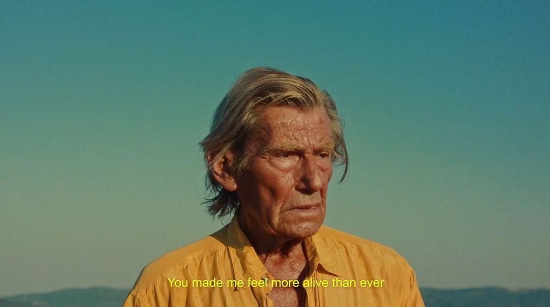 Older adult male gazes in a medium shot outdoors with a serene blue sky. He is wearing a yellow shirt, looking weathered and wise.