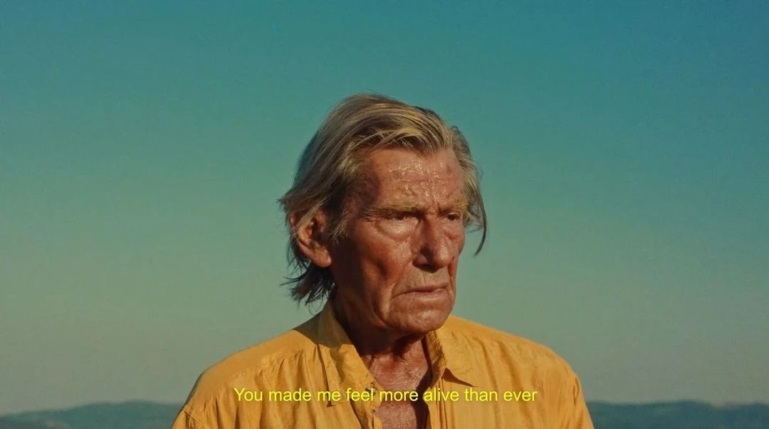 Older adult male gazes in a medium shot outdoors with a serene blue sky. He is wearing a yellow shirt, looking weathered and wise.
