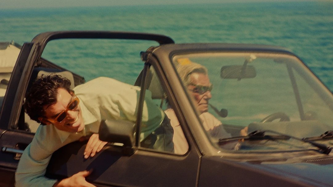 Two men ride in a vintage convertible on a sunny day near the sea. One man leans out of the car, smiling. Blue ocean in the background.