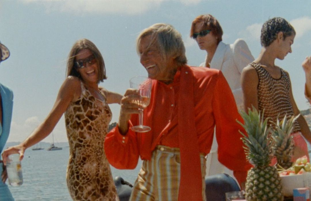 Group of people gather on a boat, laughing and drinking with pineapples and fruit, against a blue sky and water background.
