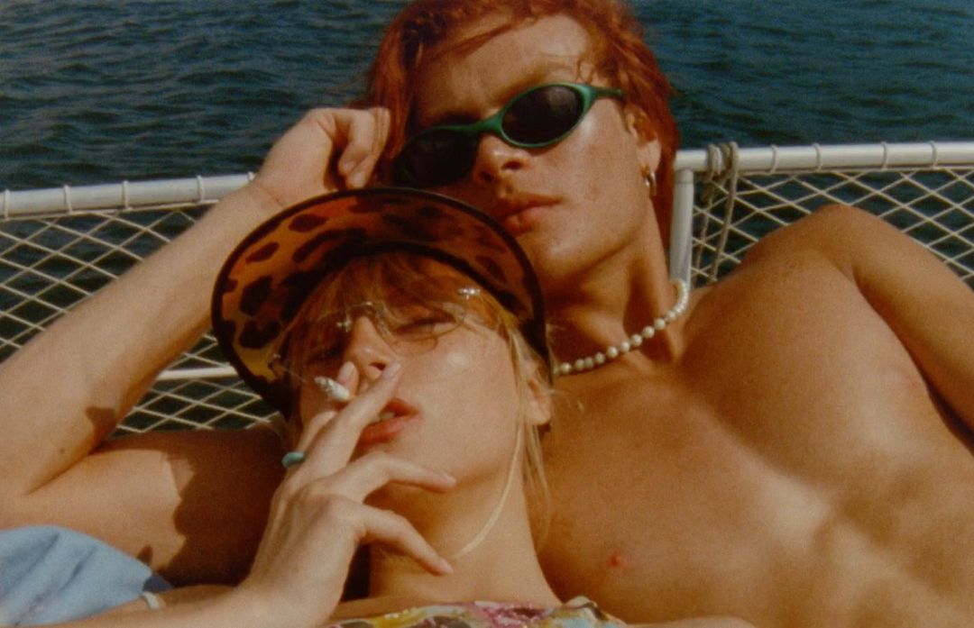 Two fair-skinned adults lounge by the ocean on a sunny day, one smoking, both wearing sunglasses, exuding a relaxed summer vibe.