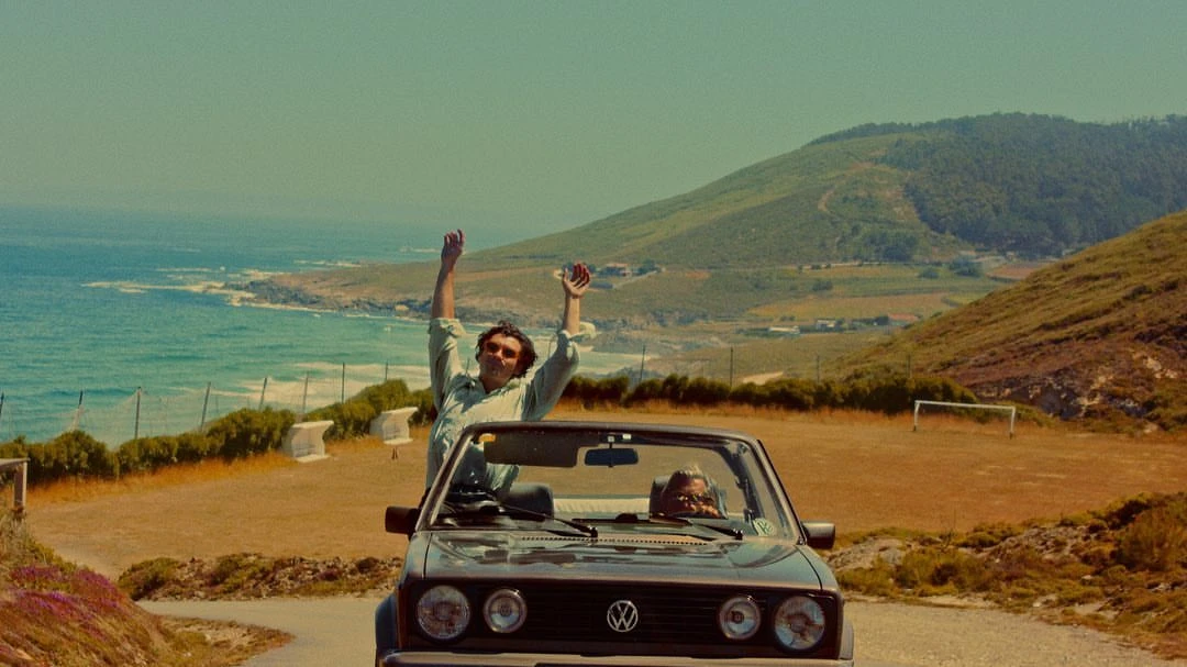 Two people drive a vintage convertible along a winding road overlooking the ocean, with a mountain visible in the background.