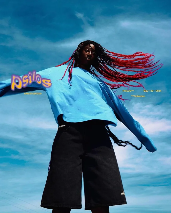 Low-angle shot of Black woman in light blue long-sleeve shirt and black shorts with red braided hair against bright blue sky by Celine Van Heel
