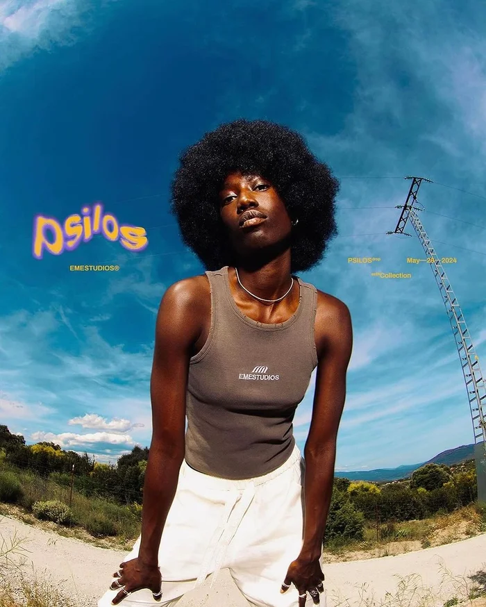 Model with an afro hairstyle wearing a grey tank top and white trousers against a bright blue sky background.