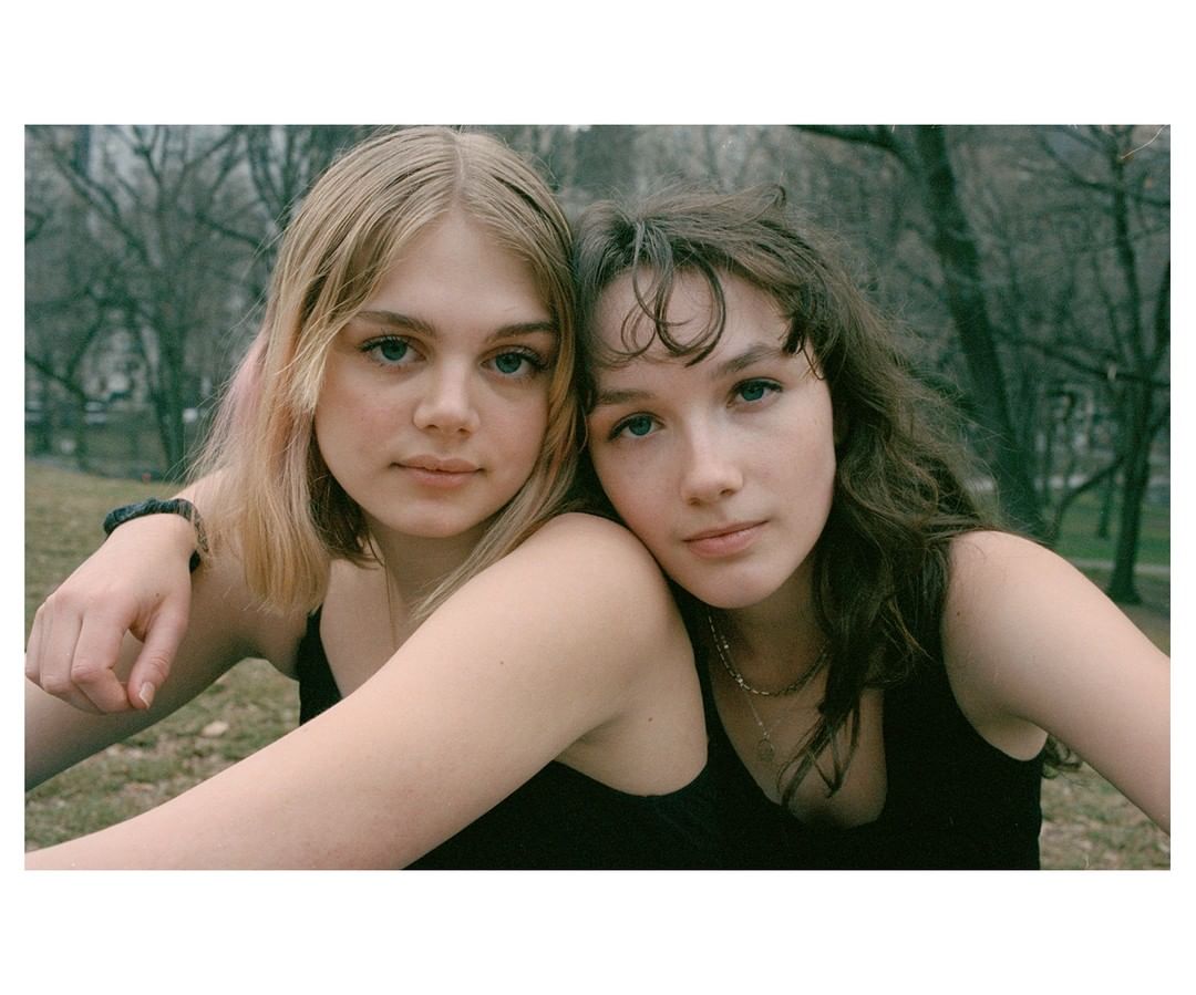 Two young women pose closely together outdoors with a blurred park and trees in the background, creating a casual, natural portrait.