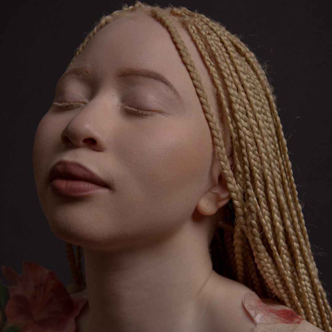 Close-up portrait of a woman with blonde braided hair and fair skin, eyes closed, with a dark grey background and red and green flowers.