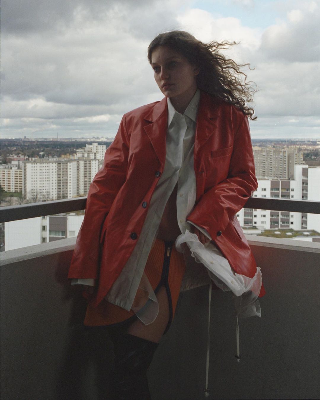 A woman with curly hair poses on a balcony in a red jacket with a city skyline and cloudy sky visible in the background, taken in natural light.