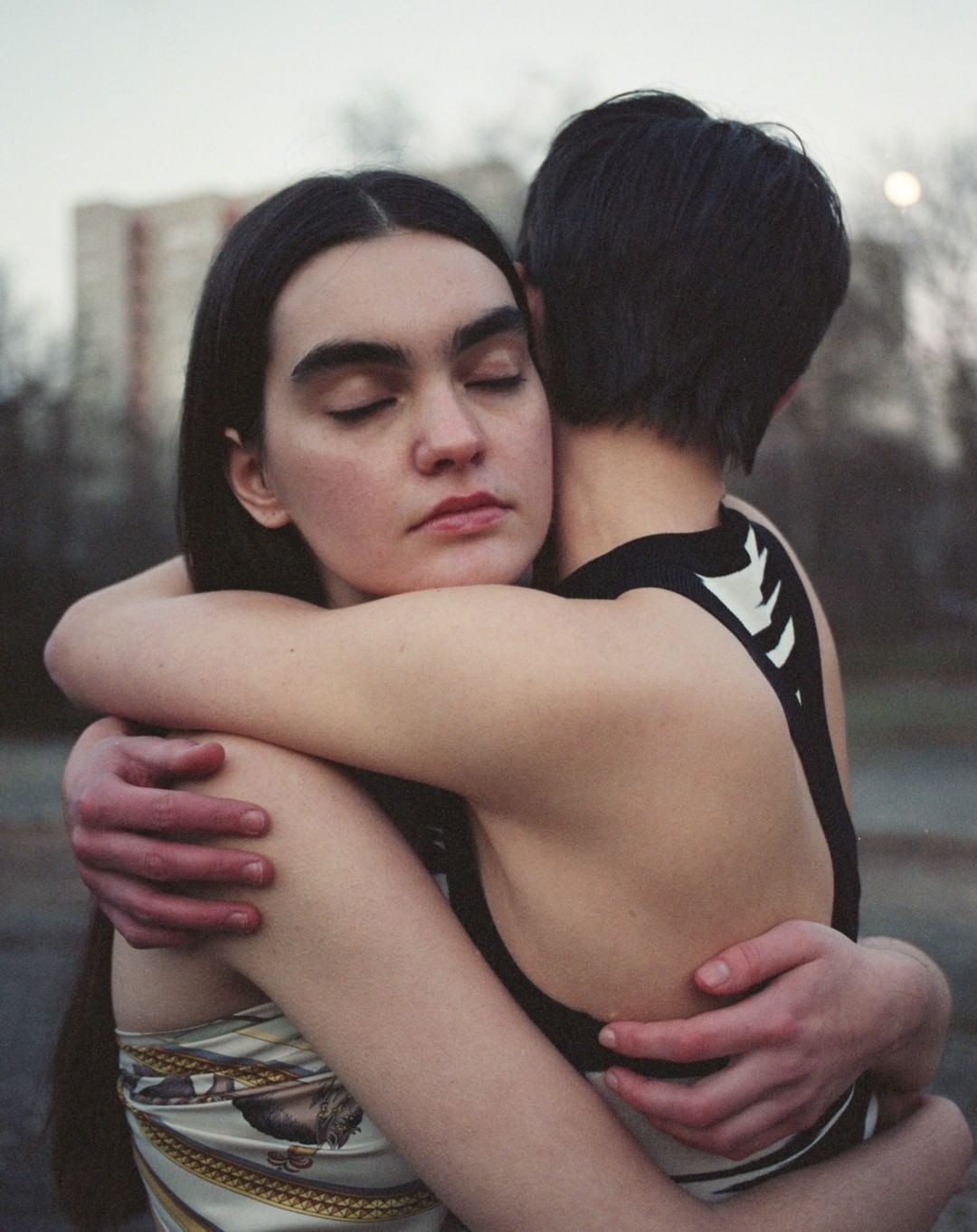 Two women embracing outdoors with neutral tones and a building in the background, portraying intimacy and connection in an urban setting.