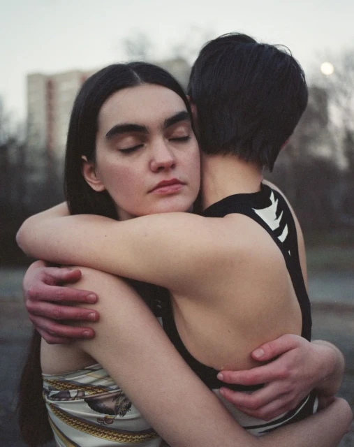 Two women embracing outdoors with neutral tones and a building in the background, portraying intimacy and connection in an urban setting.