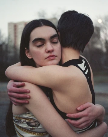 Two women embracing outdoors with neutral tones and a building in the background, portraying intimacy and connection in an urban setting.