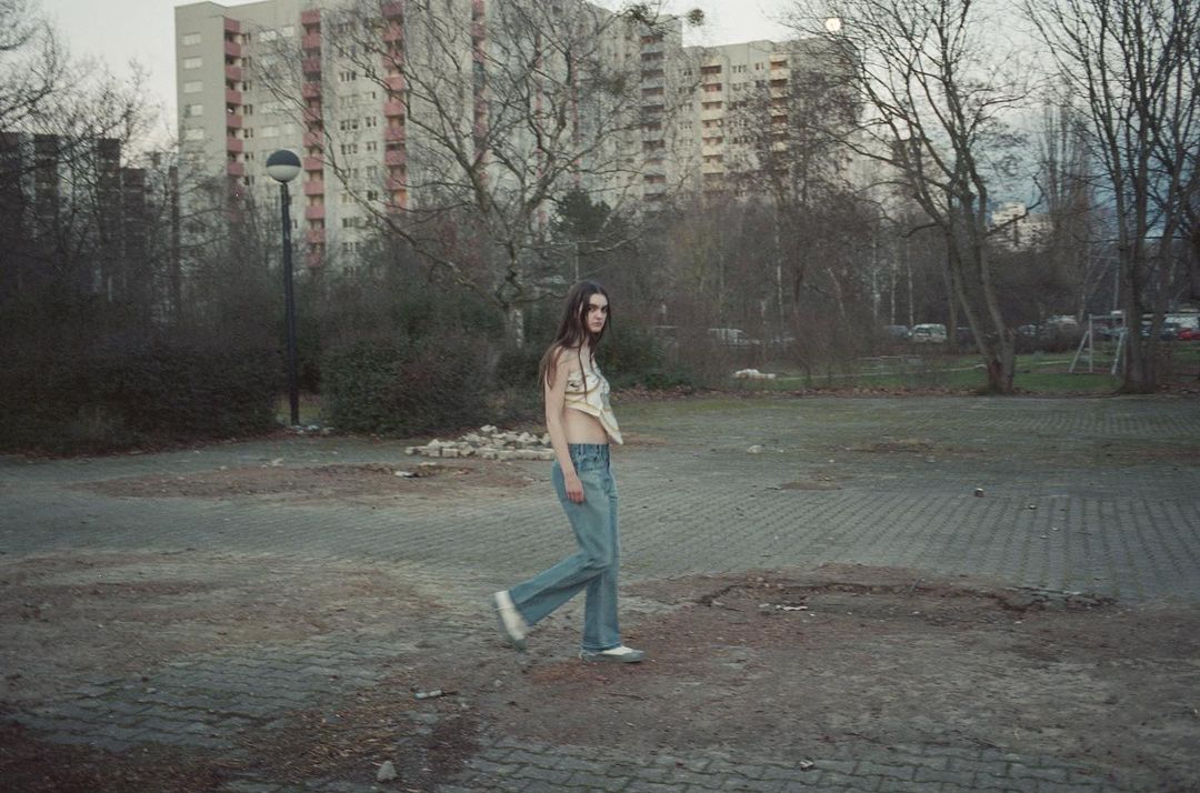 A young woman with long hair walks in an urban park on a cloudy day, wearing a cream top and light blue jeans.