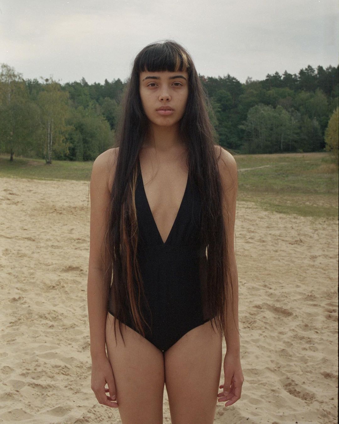 Young woman in a black swimsuit stands on a sandy beach with trees and an overcast sky in the background, captured in a medium shot.